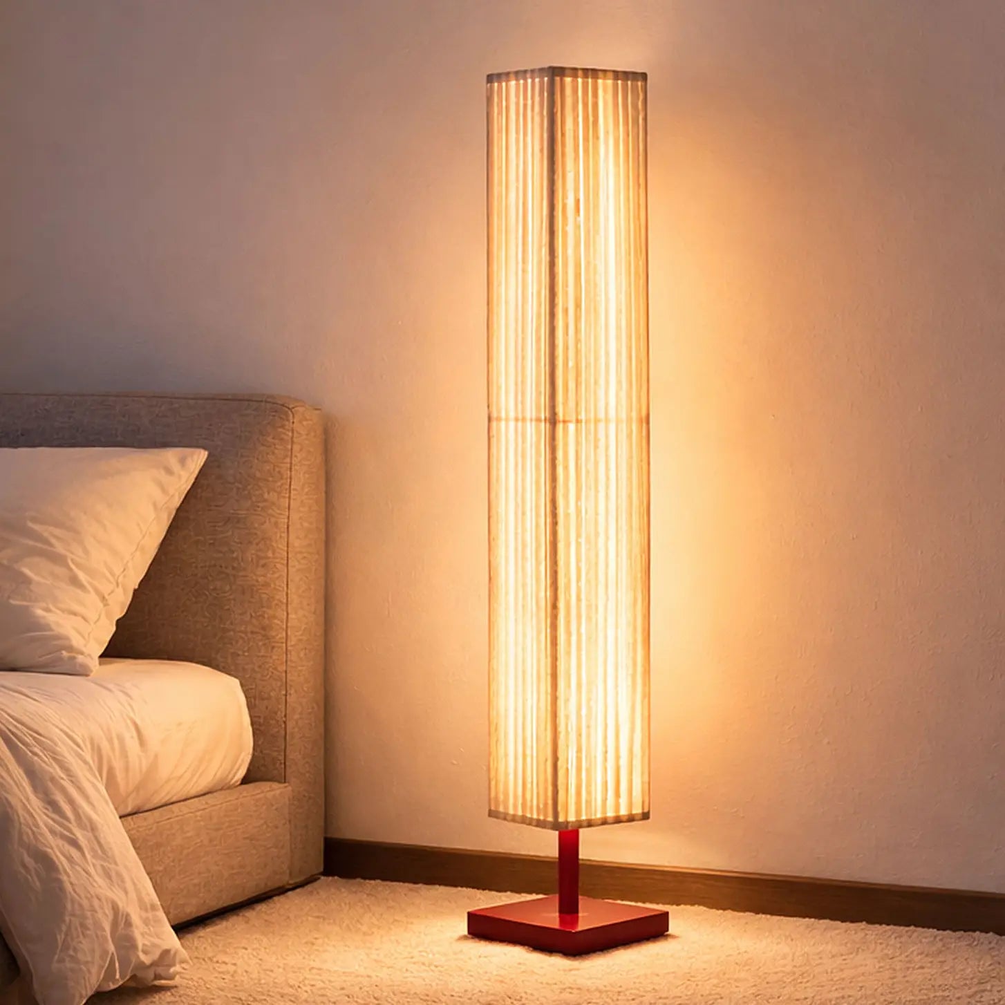 A pleated woven floor lamp lighting a bedroom at dusk